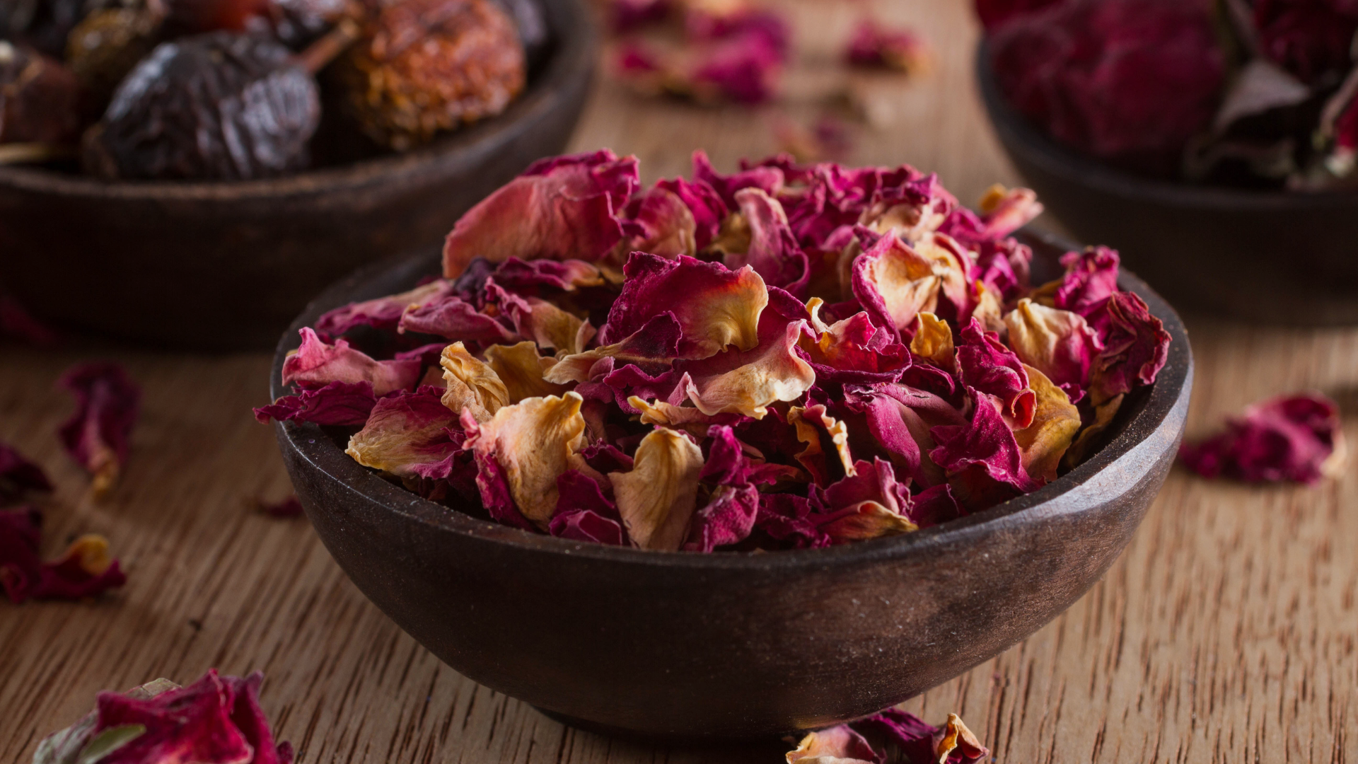 Close-up of dried rose petals in a dark wooden bowl on a rustic wood surface, used as a natural ingredient in skincare and apothecary formulations.